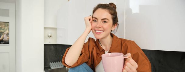 Beautiful young female model holding pink cup, sitting in kitchen and smiling at camera, woman drinking tea at home or enjoying aroma of coffee. - Photo, Image