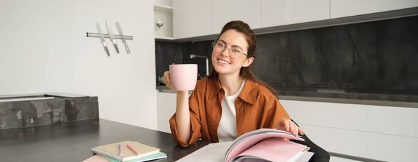 Retrato de una hermosa estudiante, mujer con gafas leyendo y bebiendo café en casa, estudiando y revisando para la lección, haciendo la tarea. - Foto, Imagen