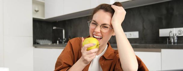 Közelkép portré aranyos fiatal nő szemüvegben, eszik alma, nevet és mosolyog, ül a konyhában. - Fotó, kép