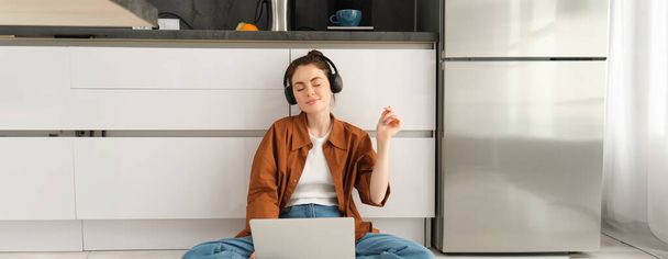 Portret van een jonge vrouw, dj die geluiden mengt op laptop, ogen sluit in een koptelefoon en naar muziek luistert, thuis op de vloer zit. - Foto, afbeelding