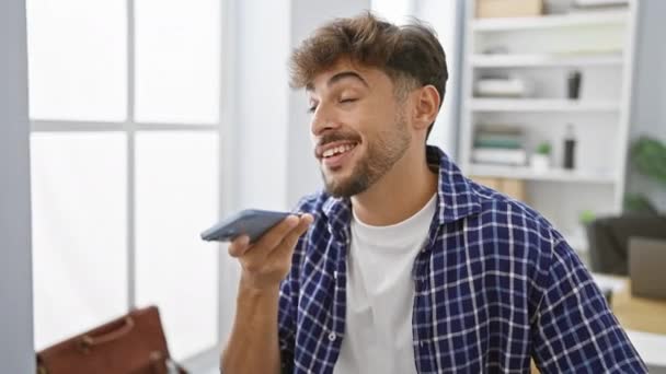Itsevarma nuori arabialainen mies hymyilee lähettäessään ääniviestin älypuhelimellaan toimistossa - Materiaali, video
