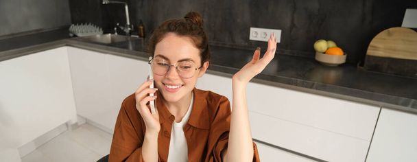 Портрет счастливой, дружелюбной молодой женщины на кухне, сидящей в очках, жестикулирующей во время разговора по мобильному телефону, отвечающей на звонок, использующей смартфон. - Фото, изображение