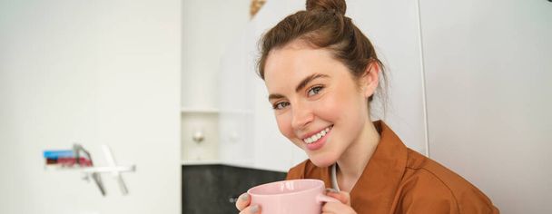 Ritratto ravvicinato di bella donna bruna con tazza di caffè, bere bevanda aromatica dalla tazza, sorridere dal piacere, seduta in cucina a casa. - Foto, immagini