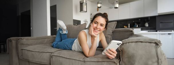 Portrét ženy, jak tráví čas doma, odpočívá na gauči v obývacím pokoji, drží smartphone, usmívá se a šťastně hledí do kamery. Koncept životního stylu - Fotografie, Obrázek