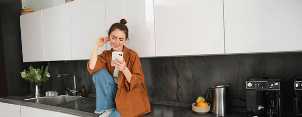 Muotokuva nuori moderni nainen istuu keittiössä laskuri älypuhelimella, chattailuun matkapuhelimella, tarkkailun sosiaalisen median sovellus ja hymyilee. - Valokuva, kuva