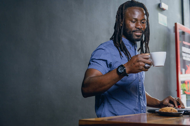 black man of African ethnicity, with dreadlocks and casual clothes, standing working with his laptop, managing his business, inside restaurant and having cup of coffee, business people concept, copy space - Photo, Image