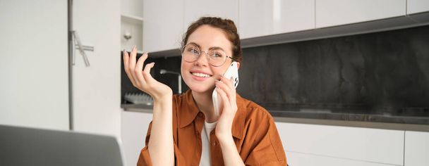 Портрет молодой женщины, предпринимателя, работающего из дома, студента, делающего телефонный звонок, сидящего на кухне с ноутбуком, разговаривающего с кем-то. - Фото, изображение