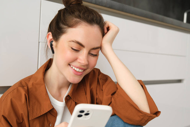 Portret van een gelukkige jonge mooie vrouw, muziek luisteren in draadloze oortelefoons, smartphone gebruiken, video 's bekijken op mobiele telefoon met hoofdtelefoon op. - Foto, afbeelding
