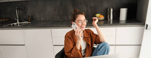 Mujer joven emocionada hablando en el teléfono móvil delante de la computadora portátil, sentado en la cocina con expresión de cara feliz, tener una conversación. - Foto, imagen