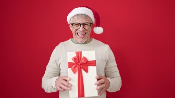 中世の灰色の髪の男性は,孤立した赤い背景の上にクリスマスの贈り物を保持する自信を持って微笑みます - 映像、動画