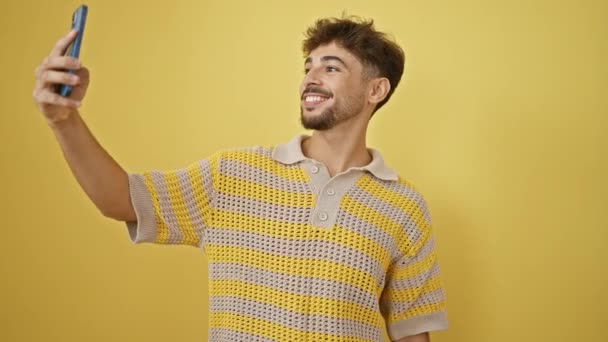 Radosny młody arabski człowiek pewnie robi fajne selfie ze swoim smartfonem na odizolowanym żółtym tle - Materiał filmowy, wideo