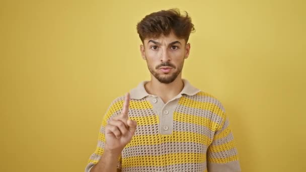 Yüzünde ciddi bir ifadeyle duran genç Arap adam, izole edilmiş sarı arka planda parmağıyla hayır diyor. - Video, Çekim