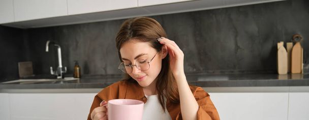 Primer plano retrato de mujer joven linda y tierna en gafas, sentado en la cocina, bebiendo té, sosteniendo la taza, se mete el pelo detrás de la oreja y sonríe. - Foto, Imagen