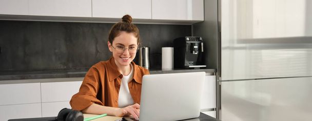 Πορτρέτο της νεαρής γυναίκας που εργάζεται από το σπίτι, δημιουργεί χώρο εργασίας στην κουζίνα, χρησιμοποιώντας φορητό υπολογιστή. Μαθητής που κάνει εργασίες στον υπολογιστή. - Φωτογραφία, εικόνα