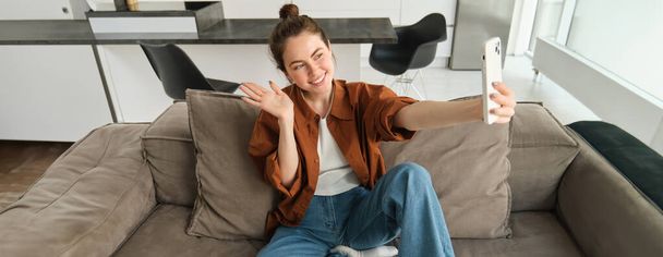 Photo of smiling young woman sitting on couch, posing on sofa in living room, taking selfies for social media. - Photo, Image