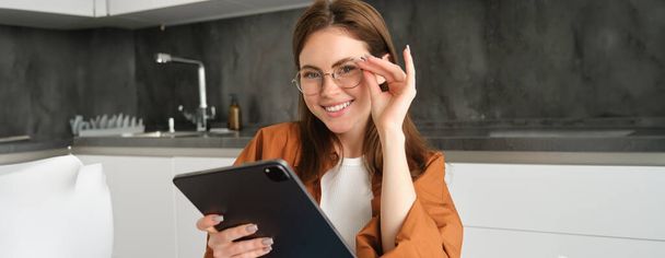 Portré magabiztos, mosolygós fiatal nő, tanuló szemüvegben, ül a konyhában, tanul a távoli, csatlakozik az online tanfolyam digitális tabletta. - Fotó, kép