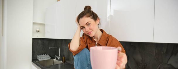Mooie jonge brunette vrouw zit in de keuken, drinkt koffie, biedt u kopje en glimlacht, ziet er gelukkig uit. - Foto, afbeelding