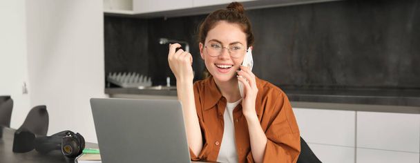 Portrait of beautiful smiling woman working from home, talking on mobile phone, calling client, self-employed businesswoman sets up workplace in kitchen, using laptop and smartphone. - Photo, Image