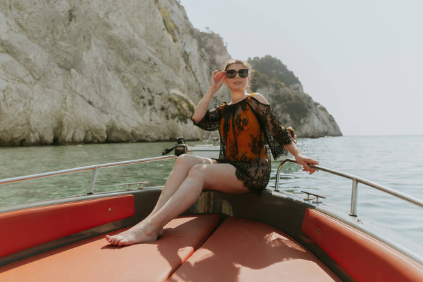 Portré egy gyönyörű fehér barnáról napszemüvegben, és egy fekete csipke köpenyről, ami a kamerába néz, és egy hajó hátulján ül, miközben a tengeren vitorlázik egy elmosódott háttér előtt. - Fotó, kép