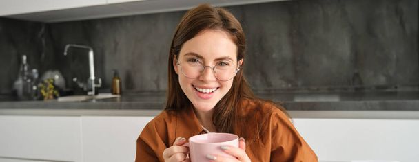 Porträt einer jungen brünetten Frau in der Küche, die sich drinnen ausruht, lächelt und Kaffee trinkt und leckeren heißen Tee genießt. - Foto, Bild