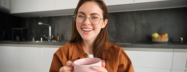Закрыть портрет красивой брюнетки в очках, отдыхающей, сидящей на кухне с чашкой чая, держащей кружку, смеющейся и улыбающейся. - Фото, изображение