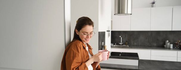 Portrait of cosy, beautiful and relaxed young woman at home, standing and leaning on wall with cup of coffee, drinking tea with thoughtful face expression. - Photo, Image