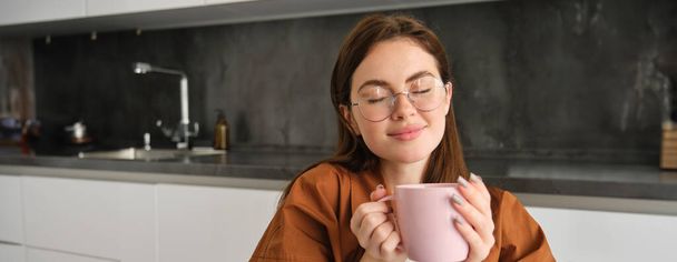 Retrato de mulher bonita em óculos, desfrutando de dia de outono acolhedor em casa, segurando xícara de café e cheira muito sabor e aroma, sorrindo satisfeito. - Foto, Imagem