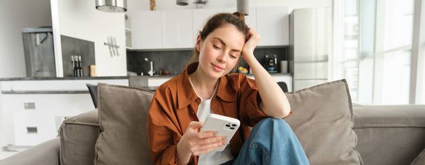 Portret van een jonge moderne vrouw die leest op mobiele telefoon, scrollende app voor sociale media op smartphone, zittend op de bank in de woonkamer in casual kleding. - Foto, afbeelding