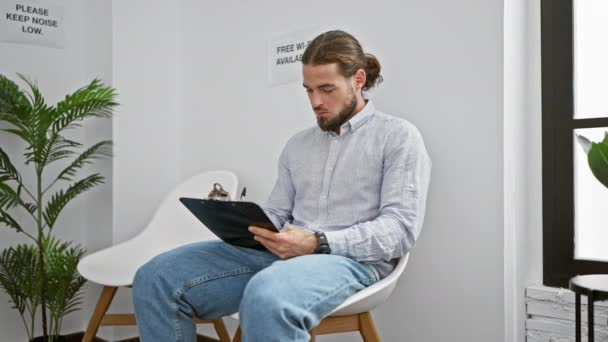 Jonge Spaanse man zit op stoel te schrijven op klembord in de wachtkamer - Video