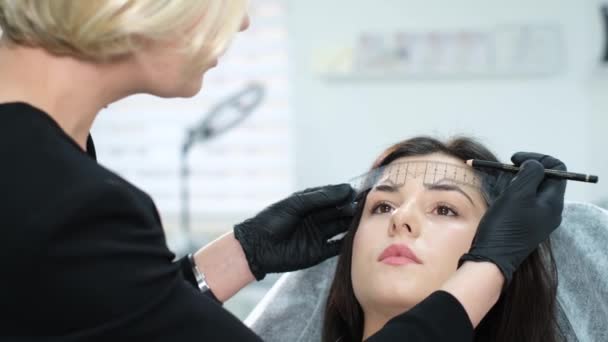 Kosmetolog měří pravítkem obočí mladé bělošky před trvalým tetováním make-upu. Tvorba permanentního make-upu obočí. - Záběry, video