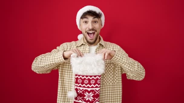 Young arab man wearing christmas hat looking inside of sock over isolated red background - Footage, Video