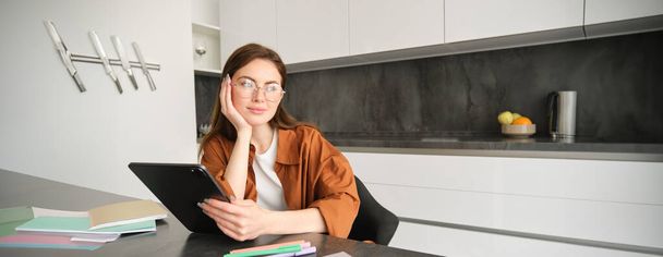 Προσωπογραφία νεαρής φοιτήτριας, γυναίκα που σπουδάζει στο σπίτι, εργάζεται εξ αποστάσεως, στήνει χώρο εργασίας στην κουζίνα της, κάθεται σε καρέκλα με ψηφιακό tablet, διαβάζει σε γυαλιά. - Φωτογραφία, εικόνα