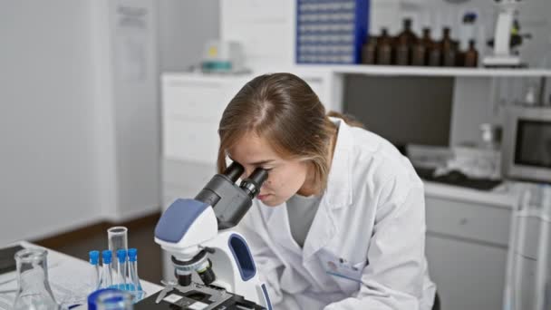 Itsevarma nuori blondi nainen tiedemies, hymyilee ristissä kädet, istuu laboratoriopöydän ääressä, työskentelee lääketieteellisen tutkimuksen mikroskoopilla vilkkaassa tiedelaboratoriossa. - Materiaali, video