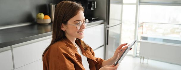 Vista lateral da jovem mulher de óculos, linda menina lendo no tablet digital, mensagens, compras on-line com gadget, sorrindo, sentado na cozinha em casa. - Foto, Imagem