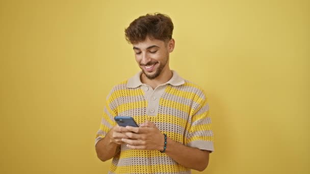 Handsome young arab man, confidently texting on his smartphone, smiling joyfully against a vibrant isolated yellow background - Footage, Video