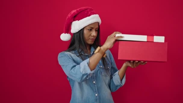 Afroamerikanerin trägt Weihnachtsmütze beim Auspacken von Geschenk und wirkt aufgebracht über isolierten roten Hintergrund - Filmmaterial, Video