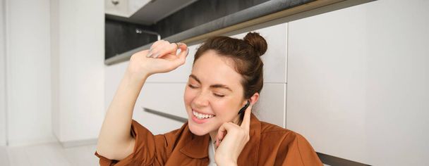 Portrait of young carefree woman sits on floor at home, dances while listens to music in wireless headphones, singing along favourite song. - Photo, Image