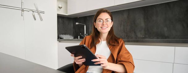 Elektronikus tanulás és távoli munkahelyi koncepció. Szemüveges fiatal nő, otthon tanul a konyhában, digitális táblagépen olvas, megnézi a projektjét, kapcsolódik az online találkozóhoz. - Fotó, kép