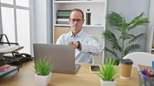 Hombre hispano de mediana edad, un trabajador de negocios serio y exitoso, sentado en la mesa de su oficina, concentrándose en su trabajo, mirando al reloj, usando un teléfono inteligente, con gafas puestas, en interiores. - Metraje, vídeo