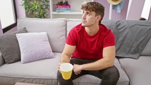 Otthon, egy magabiztos fiatal spanyol férfi élvezi a reggeli csésze kávét, kényelmesen ellazult a kanapén. jóképű arckifejezése pozitív és örömteli a fertőző, szívmelengető mosolyából.. - Felvétel, videó