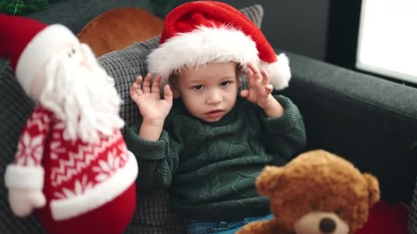 Adorable niño rubio sentado en el sofá con sombrero de Navidad en casa - Imágenes, Vídeo