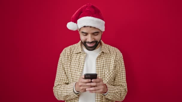 Junger hispanischer Mann mit Weihnachtsmütze und Smartphone mit Siegerausdruck vor isoliertem roten Hintergrund - Filmmaterial, Video