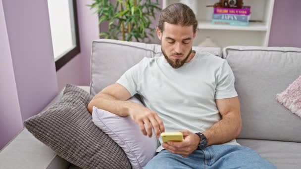 Joven hombre hispano usando smartphone sentado en sofá sufriendo de dolor cervical en casa - Imágenes, Vídeo