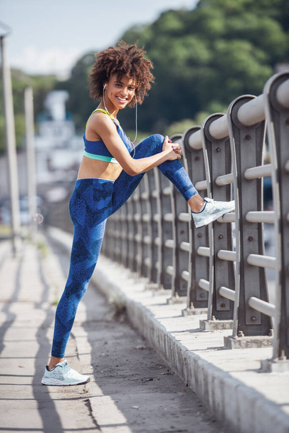 Beautiful girl in sportswear and earphones is looking at camera and smiling while stretching before morning run - Photo, Image