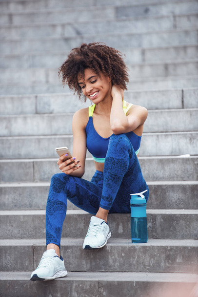Gyönyörű lány sportruházatban használ egy okos telefont, és mosolyog, miközben pihen a lépcsőn reggeli futás közben - Fotó, kép