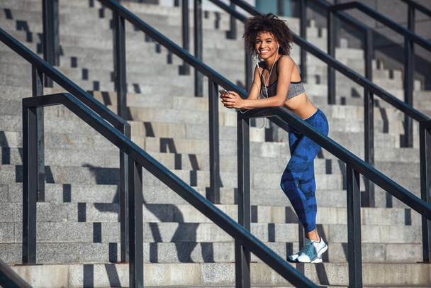 Beautiful girl in sportswear and earphones is looking at camera and smiling while resting on stairs during morning run - Photo, Image