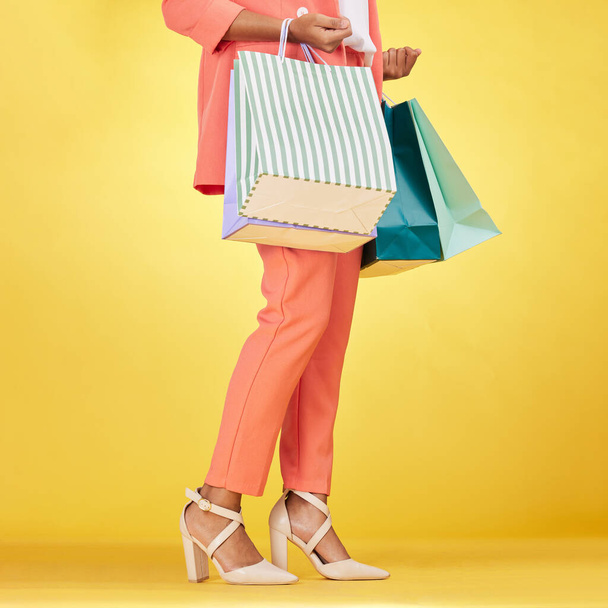 Legs, woman and shopping bag in studio for fashion, retail deal and financial freedom on yellow background. Closeup of customer, gift bags and discount from clothes store, shoes sales and promotion. - Photo, Image