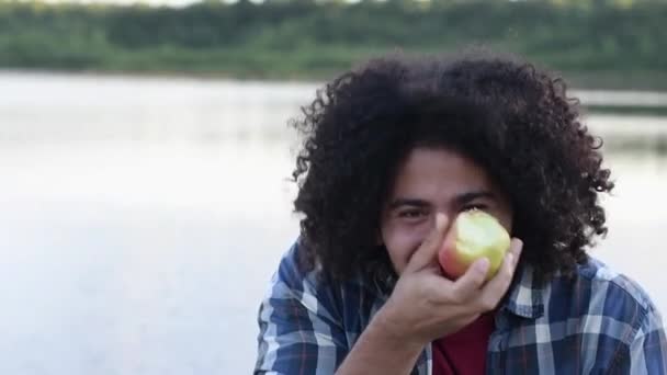 heureux cheveux noirs bouclés beau mec arabe étonné mord une pomme et rit gaiement, montre ses dents blanches droites, regarde la caméra, des images FullHD de haute qualité - Séquence, vidéo