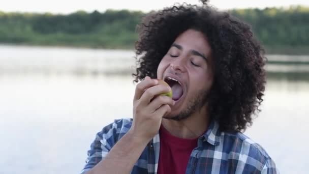 gelukkig zwart harig krullend knap verbaasd Arabisch guy eten groene appel in de natuur tijdens een wandeling en lacht vrolijk, kijkt naar de camera, tegen de achtergrond van een meer, Hoge kwaliteit FullHD beeldmateriaal - Video