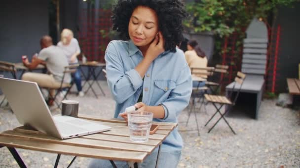 Fröhliche Afroamerikanerin mit Kopfhörern, während sie ihr Smartphone in der Hand hält. . Junge Frauen hören Musik, während sie am Laptop im Café arbeiten. Einsatz moderner Technik im Sitzen auf der Terrasse - Filmmaterial, Video
